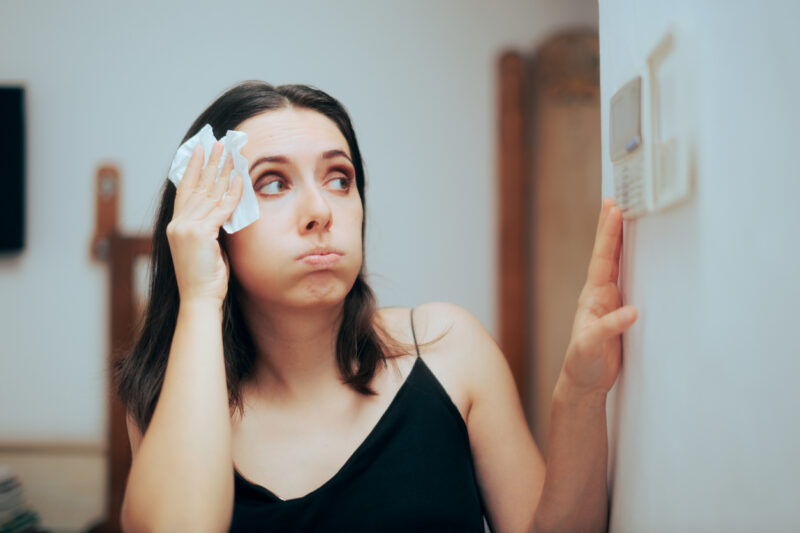 Woman adjusting AC temperature on a home thermometer while wiping sweat from her brow